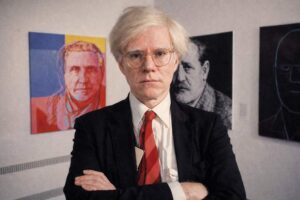 Andy Warhol - Porträtfoto silber-weißen Perücke, Brille und roter Krawatte vor seinen Kunstwerken