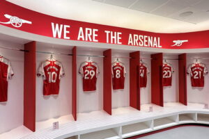 Arsenal Museum - London - Ausstellung: Spielerkabine mit roten Trennwänden, weißen Sitzbänken und den Rückansichten der Trikots von Havertz, Ødegaard und Rice unter dem geschwungenen Schriftzug WE ARE THE ARSENAL