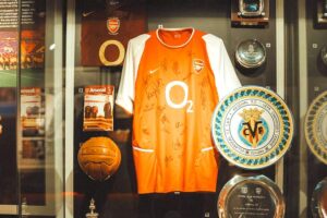Arsenal Museum - London - Ausstellung: Eine gläserne Museumsvitrine mit einem von der Mannschaft signierten, orange-weißen O2-Trikot, einem alten Lederfußball und feierlichen Gedenktellern zum Champions-League-Finale 2006 gegen Barcelona