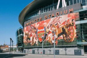 Arsenal Museum - London - Emirates Stadium - Außenansicht: Die geschwungene Glas- und Stahlfassade des Stadions mit dem EMIRATES STADIUM Schriftzug und der riesigen Wandmalerei mit dem Arsenal-Vereinswappen und historischen Kanonen