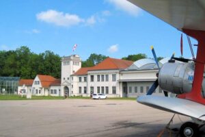 Deutsches Museum Flugwerft Schleißheim - Außenansicht mit dem historischen Gebäude, dem Kontrollturm und einem Propellerflugzeug im Vordergrund