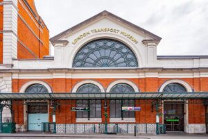 London Transport Museum - London - Außenansicht: Die historische rote Backsteinfassade des Museumsgebäudes am Covent Garden mit einem großen, verzierten Bogenfenster und dem goldenen Schriftzug - LONDON TRANSPORT MUSEUM - auf dem weißen Giebel