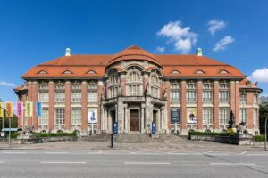 Museum am Rothenbaum - Hamburg - Außenansicht: Ein großes, symmetrisches Gebäude mit rotem Ziegeldach und bunten Fahnen mit der Abkürzung MARKK vor dem Eingangsbereich