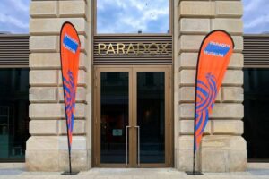 Paradox Museum - Hamburg - Eingangsbereich: Der moderne Eingangsbereich mit zwei orange-blauen Werbefahnen vor einer klassischen Steinfassade und einer Bronzetür