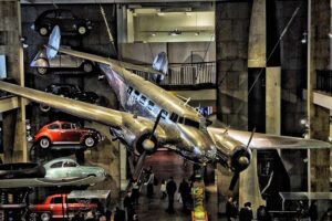 Science Museum - London - Ausstellung: Blick in die Halle - Making the Modern World - mit einer im Vordergrund aufgehängten, silbernen Lockheed 10A Electra aus dem Jahr 1935 und historischen Autos an der Wand