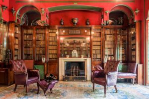 Sir John Soane’s Museum - London - Library Dining Room: Historische Bibliothek und Speisezimmer mit roten Wänden, dunklen Holzregalen und zwei Ledersesseln vor einem Kamin