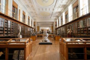 The British Museum - London - Kings Library: Ein langer, prachtvoller Saal mit historischen Bücherschränken hinter Glas, die sich über zwei Ebenen erstrecken, und antiken Büsten auf Podesten