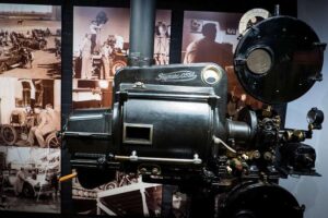 The Cinema Museum - London - Filmprojektor: Ein schwarzer Vintage-Filmprojektor von OSSA aus den 1950er-Jahren vor einer Wand mit historischen Schwarz-Weiß-Fotografien