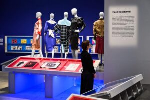 The Design Museum - London - Ausstellung Blitz The Club that Shaped the 80s: Schaufensterpuppen präsentieren farbenfrohe Mode der 80er Jahre vor einer blauen Wand mit Informationstafeln