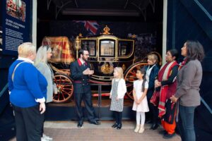 The Royal Mews - London - Führung: Ein Guide im Royal Mews führt eine Besuchergruppe vor der Queen Alexandra’s State Coach durch die Ausstellung